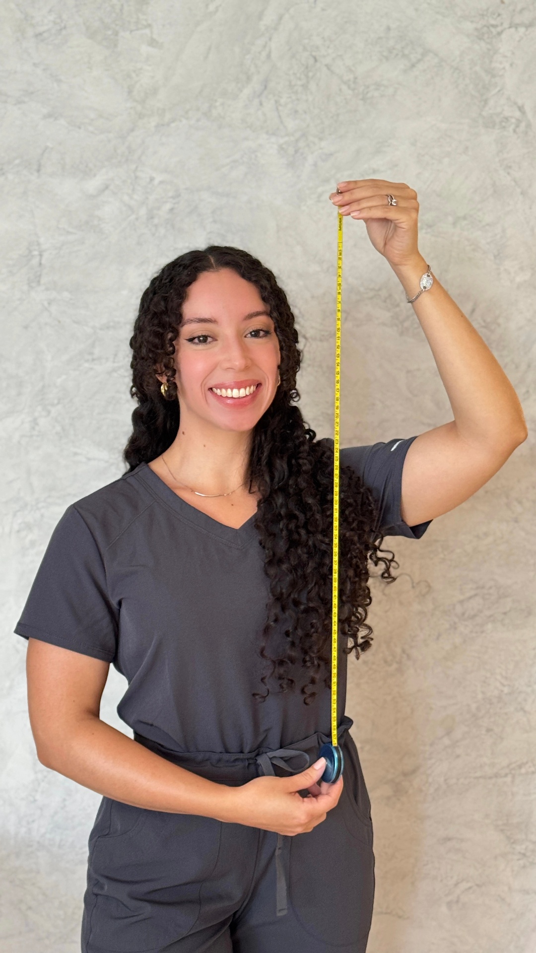 Megan Maldonado de pie sonriendo con uniforme gris, sosteniendo una cinta métrica amarilla extendida en vertical